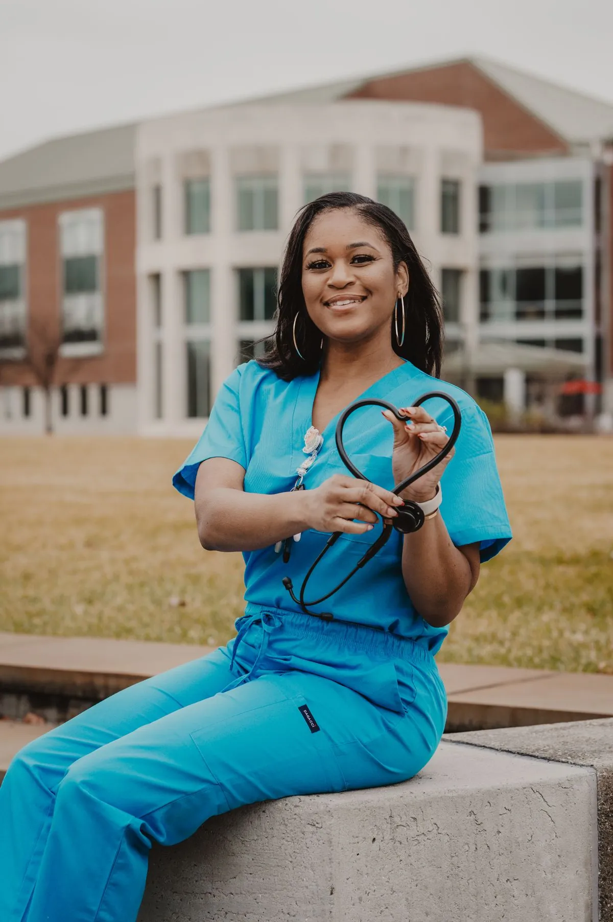 A nurse in teal scrubs holding a stethoscope outside a hospital